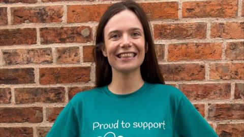 A woman with long brown hair smiles while standing in front of a brick wall. She is wearing a green t-shirt which says "proud to support" on it.