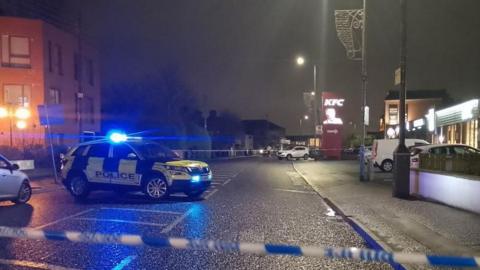 A road with a blue and white police tape, there is a yellow and blue police jeep and red KFC sign in the background.