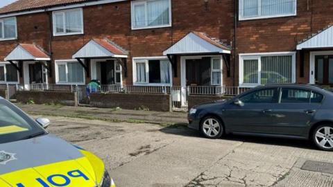 Two cars - a grey vehicle and a PSNI car - parked outside a row of red brick houses in north Belfast