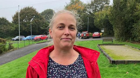 Karen Bennett stood on the green of the village hall. She is wearing a floral blouse and a red jacket. She is looking at the camera with her hair tied back. Multiple cars are parked in the background. 