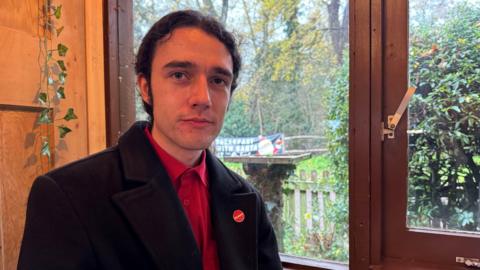 A man wearing a black jacket and a red shirt, wearing a badge that says 'Labour'. He is sat down inside a cafe, with a window behind him showing  a green lawn. 