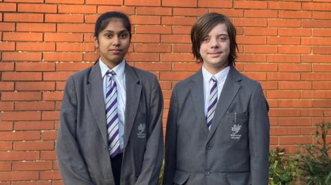 Students Nathan and Gargi looking at the camera. They are wearing school uniform made up a grey blazer and purple tie. They are standing in front of a brick wall and there are some flowers in the background. 