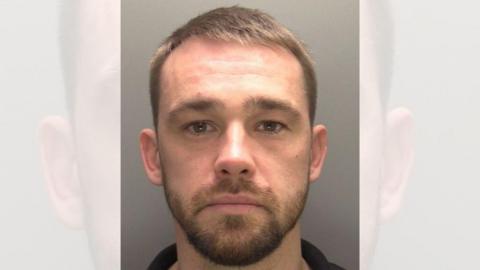 A police custody photo of a young man with short cropped brown hair and a brown beard. His eyes are bloodshot in the photo.