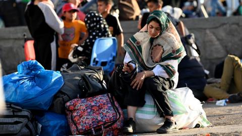 A woman sits with a pile of bags containing her belongings at the border crossing between Iran and Afghanistan, with other refugees visible in the background.