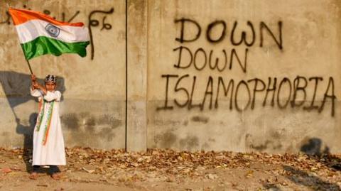 A Muslim child seen holding the Indian flag next to a wall that says 'Down down Islamophobia'