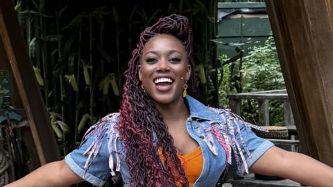 Georgina Onuorah in costume as Lulu in Shucked at Regent's Park Open Air Theatre. She is standing near the stage and rows of fake corn are behind her. She is wearing an orange T-shirt with a denim jacket/waistcoast over the top. She is looking directly at the camera and is smiling with her mouth wide open.