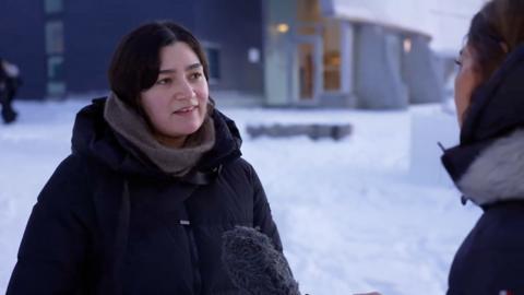 Woman with black hair, wearing a black jacket, being interviewed in the snow.