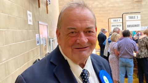 Conservative Councillor Adrian Hardman is smiling at the camera, wearing a blue suit and a blue Conservative rosette.