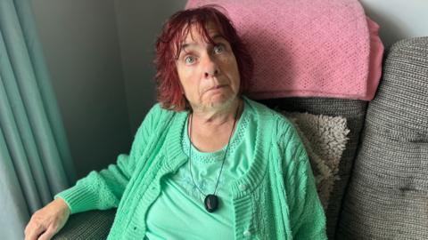 Woman in green top and cardigan with short red hair sat down on sofa 