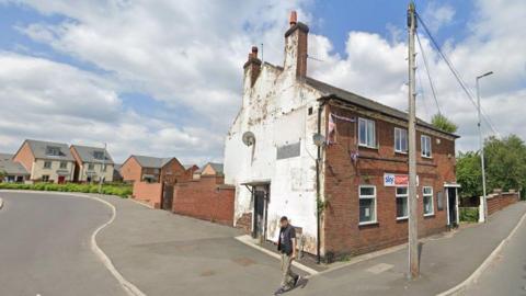 A man walks past a dilapidated building on a sunny day. The two-storey building has peeling white paintwork on a side wall, and the remains of a frayed Union Jack flag on a front wall. There are residential houses in the background.