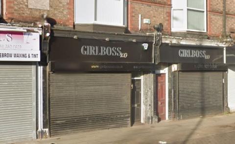 Girlboss business in Cressington. It has a black sign reading Girl Boss, with the shop's shutters down. A black door is the entrance - next to a red door - with windows above. On a street next to a nail salon advertising also waxing and tinting. 