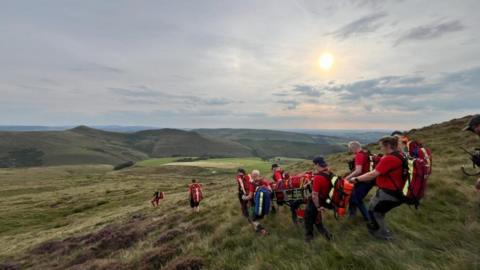 A mountain rescue team wearing red shirts and carrying backpacks use a wheeled stretcher to transport a woman down a mountain