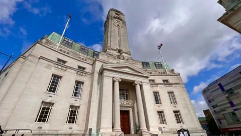 Luton's four-storey neo-classical Town Hall built in white stone with tall columns either side of the entrance. There are narrow windows on either side of the entrance, and a tall clock tower on the roof. There are two flagpoles on either side of the roof.