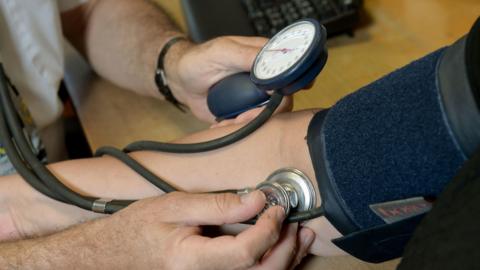 A doctor is using a stethoscope and blood pressure monitor on the arm of a patient. The image is a close-up of the patient's forearm and the doctor's hands.