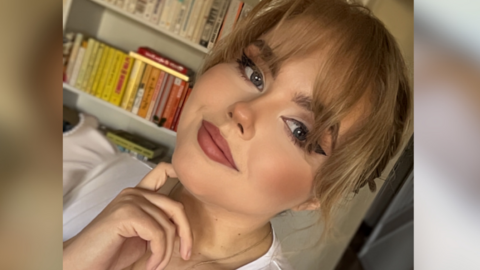 Anya Roberts stands in front of a bookshelf. She has blonde hair tied back, with a long fringe, and wears a white t-shirt with a silver necklace.
