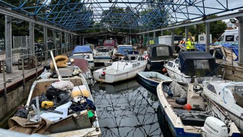 A narrow body of water with many boats in it, some full of rubbish and none in a very good state of repair. Above them is a roof structure made of blue poles and the waterway is surrounded by metal railings and concrete walls.