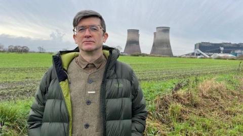 Professor Richard Brook has short hair and is wearing a green jacket over a tweed jacket and is standing in front of the Fiddler's Ferry cooling towers