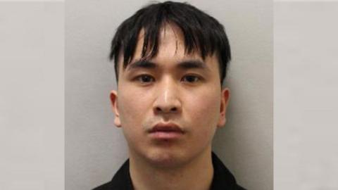 Mugshot of Zou in front of a plain wall looking stern faced