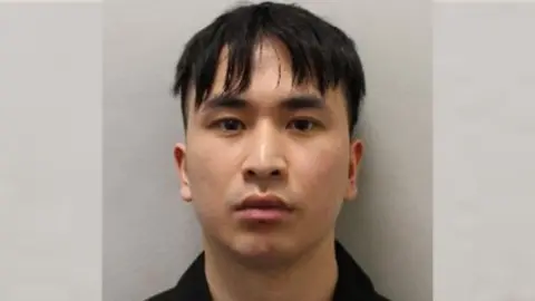 Mugshot of Zou in front of a plain wall looking stern faced