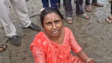 A woman wearing an orange saree sitting on wet sand with the feet of people standing behind in the frame 