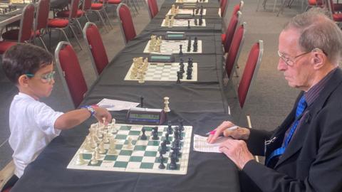 A young boy wearing a white shirt and colourful glasses, with short dark hair sits on a red chair at a table opposite a man with grey hair, wearing a suit and glasses, with a newspaper rolled up in his pocket. They are playing chess. There are many other chess boards and chairs lined up along tables in the room.