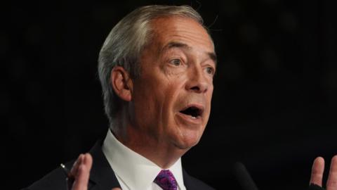 Reform UK leader Nigel Farage speaks in front of a black background while wearing a white shirt, black suit and purple tie.