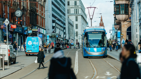 A blue tram travels down Corporation Street in Birmingham in the day with shoppers walking up and down the street past the shops. There is a van parked on the left-hand side of the street