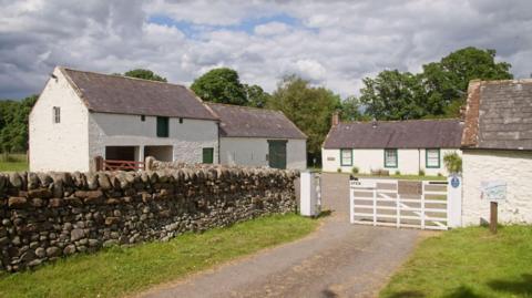 White farm buildings surrounded by a stone wall. Trees are in the background and there is a white fence and gate at the entrance.