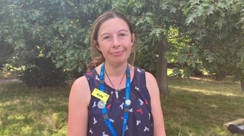 Kate Monfort from the Recovery College. Kate is stood in Stoke Park, Guildford. Is is wearing a lanyard and a navy vest top. 