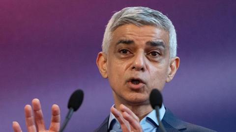 Sadiq Khan is pictured from the shoulders up giving a speech. Two microphones are visible in the foreground and he stands in front of a blurred purple background.