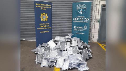 Multiple silver parcels are piled up on the ground in front of two blue signs, one for the Garda National Drugs and Organised Crime Bureau and the other for Irish Revenue. 