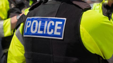 A police officer seen from behind, wearing a high-vis jacket, black vest and a large badge with the word POLICE on the back. 