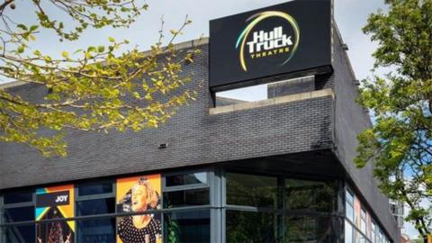 Outside of Hull Truck Theatre which is built of black bricks with posters in the ground floor windows and a sign with the theatre's name on it at the top. There are tree branches in the foreground.