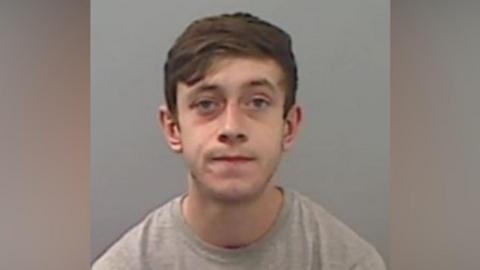 Police mugshot of Harrison Simpson. He is a young man with short brown hair swept across his forehead and is slightly grimacing, wearing a grey t-shirt.