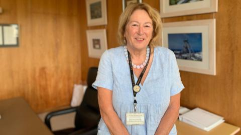 Councillor Linda Taylor is smiling at the camera as she stands in an office. The wooden walls have many framed pictures and photos on them and part of a desk and chair can be seen. The councillor is wearing a pale blue dress, a matching chunky bead necklace and a lanyard which reads CORNWALL COUNCIL.