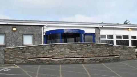 The front of Ramsey Cottage Hospital, which is a stone building that has a blue-framed glass revolving door with a stone wall in front of it.