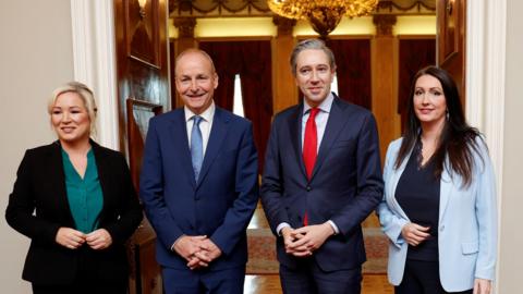 Four people stood smiling. Leftmost is a woman with blonde hair, wearing a black suit jacket and trousers, and a green shirt. On her right is a man wearing a navy suit, with a white shirt and light blue tie. On his right is a man wearing a navy suit with a white shirt and a red tie. Rightmost is a woman with darl hair wearing a black top and trousers and a light blue suit jacket. They are standing in front of large wooden doors, which are open. 