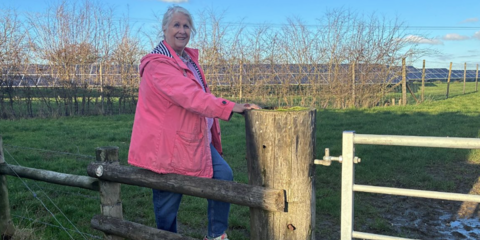 Woman in pink coat and blue jeans sitting on wooden fence near field.
