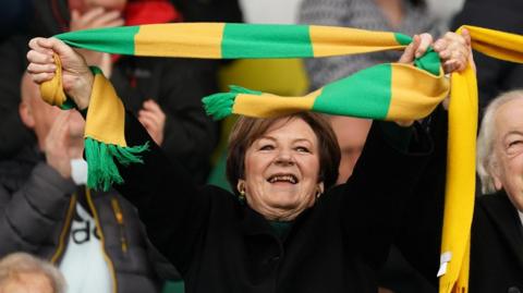 Delia Smith in a crowd at a Norwich City football match. She is holding a yellow and green striped scarf above her head with her mouth open while enjoying the moment. She has short brown hair and is wearing a black coat.