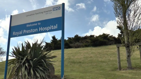A blue and white Royal Preston Hospital sign on a grassy verge. There are trees behind it.