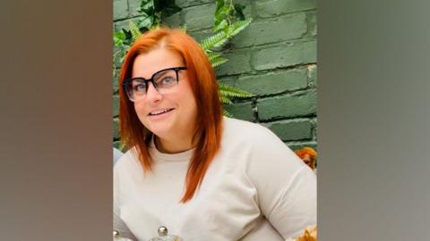 Sian Ashcroft smiles at the camera. She has bright ginger shoulder-length hair. She wears dark-rimmed glasses and a cream t-shirt.