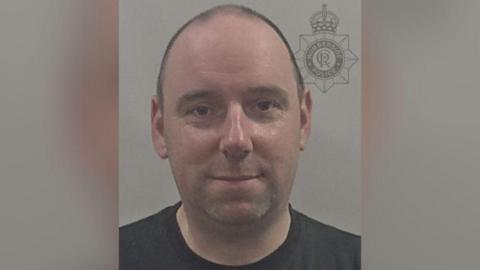 A man with very short hair which is bald at the front is smiling for a police photo. He is wearing a black t-shirt and the background is grey.