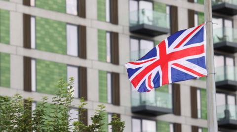 A union jack flying in front of a green-clad tower block