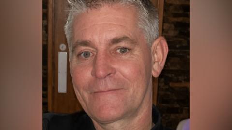 Close-up headshot of a man with short grey hair, looking toward the camera. The image is tightly cropped around the face, with a wooden interior background visible behind. The person is wearing a dark top and is photographed indoors under warm lighting.