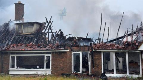 The burnt out remnants of a bungalow. Its roof has been destroyed with only a few frayed wooden beams remaining and set against thick grey smoke rising from the property. Many of the windows of the bungalow have shattered as a result of the intense heat from the fire. A lone firefighter on an aerial appliance is faintly visible on the other side of the property, through the smoke.