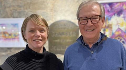 Alice Gill and Peter Neville are smiling while standing next to each other. Alice Gill has blonde bobbed hair and is wearing a black roll neck jumper. Peter Neville is wearing glasses and a blue shirt with a blue sweatshirt on top. A blurred sign saying Guernsey Chamber of Commerce can be seen behind them.