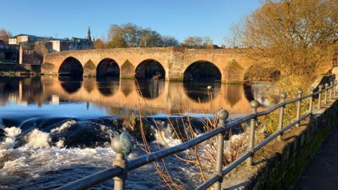 A view of Devorgilla Bridge at Whitesands, Dumfries, on a crisp, winter morning. The water under the bridge is almost still and there is a metal fence in the foreground.