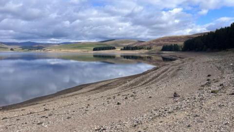 A reservoir with a stony beach in the foreground and hills in the background