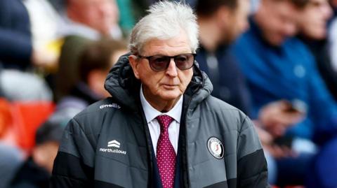 Roy Hodgson with his hands in his coat pockets and wearing sunglasses, watches Bristol City play
