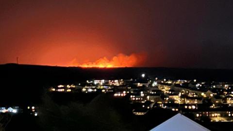 Flames from a fire on Rosenannon Downs seen from Wadebridge during the night. Dozens of houses can be seen in the foreground with their lights on. Flames and smokes are going up into the night sky with an orange glow around the site.